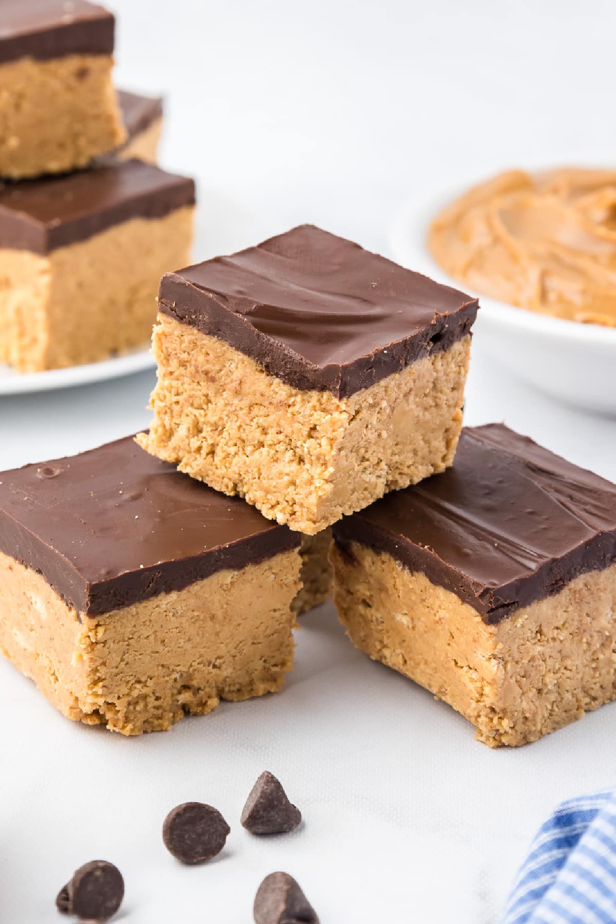 Three peanut butter squares topped with a layer of chocolate stacked with more stacked on a plate int he background on a counter.