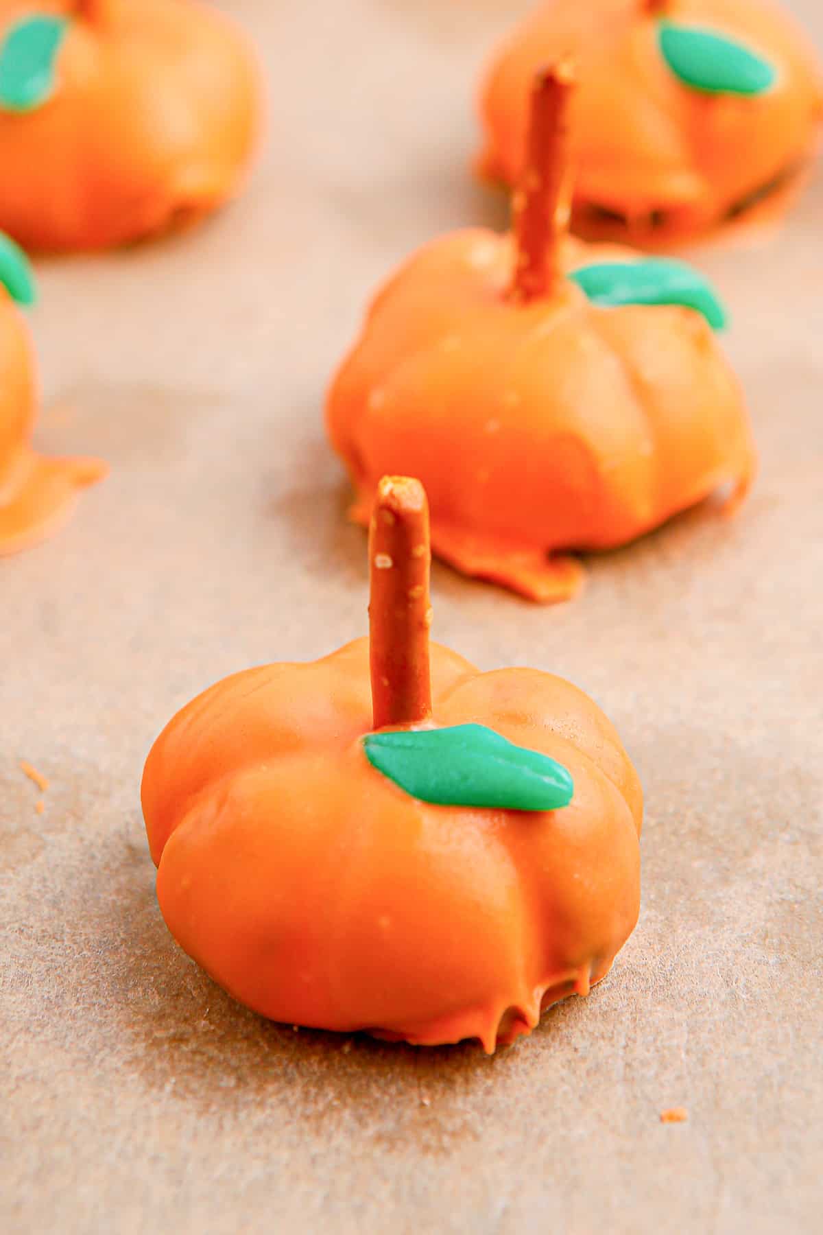 Pumpkin shaped Oreo ball with a candy leaf and pretzel stem on parchment paper with more on the pan in the background.