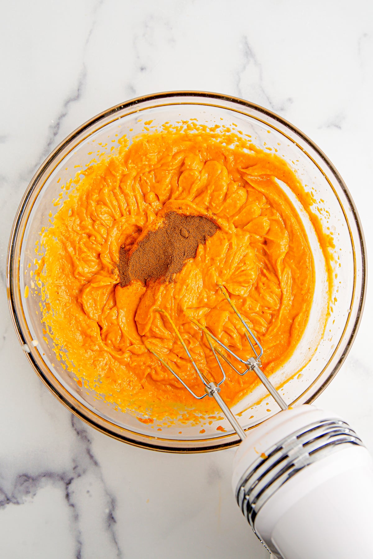 A glass bowl with an electric mixer whipping an orange pumpkin pie filling being mixed with pumpkin pie spice from overhead.
