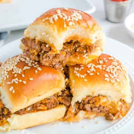 Sloppy Joe sliders stacked on a plate with sesame seeds and the top sandwich is missing a bite close up from the side.