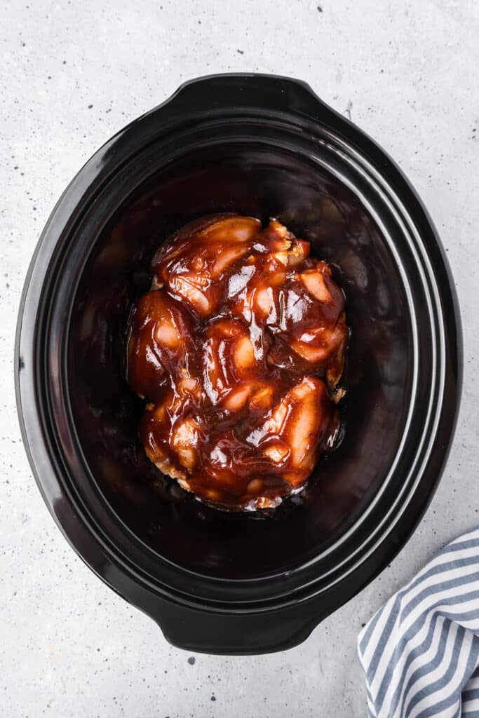 Raw boneless chicken thighs in a slow cooker base covered in bbq sauce from above.