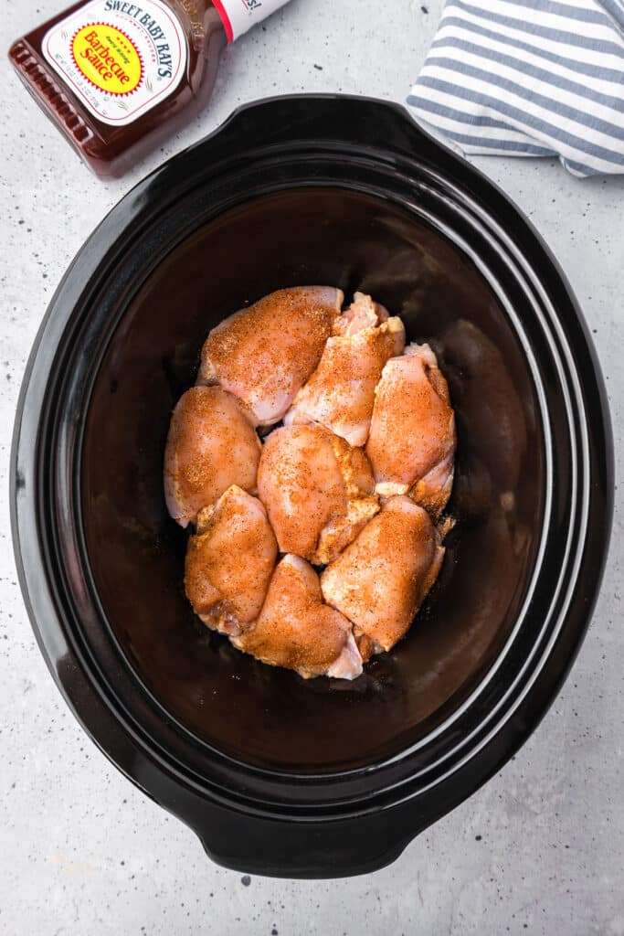 Raw spiced chicken thighs in the slow cooker from above with a bottle of bbq sauce on the counter nearby.