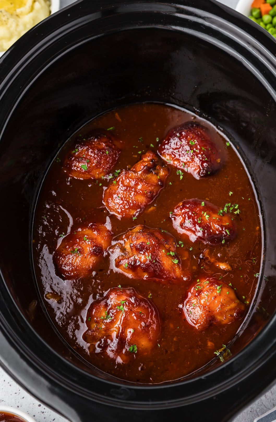 Boneless chicken thighs cooked in bbq sauce close up in a slow cooker from above with other bowls of vegetable next to the crockpot nearby.