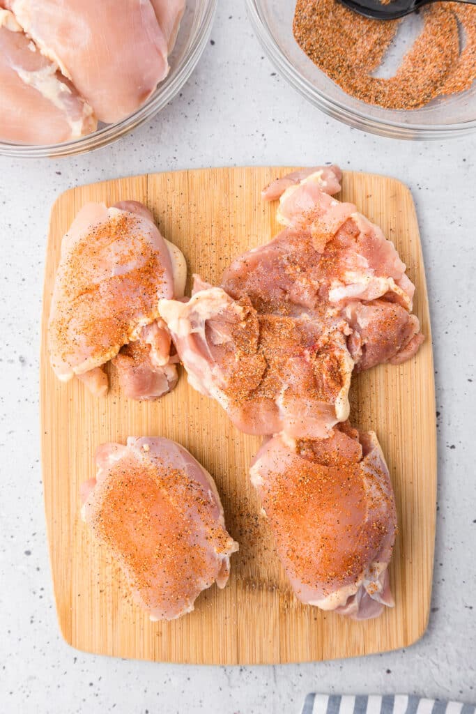 Raw boneless chicken thighs on a cutting board from above being rubbed with spices with one chicken thigh flipped over to the back.