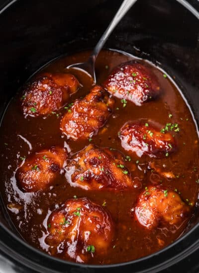 Square view of bbq chicken thighs in bbq sauce in a slow cooker with a spoon resting in the sauce.