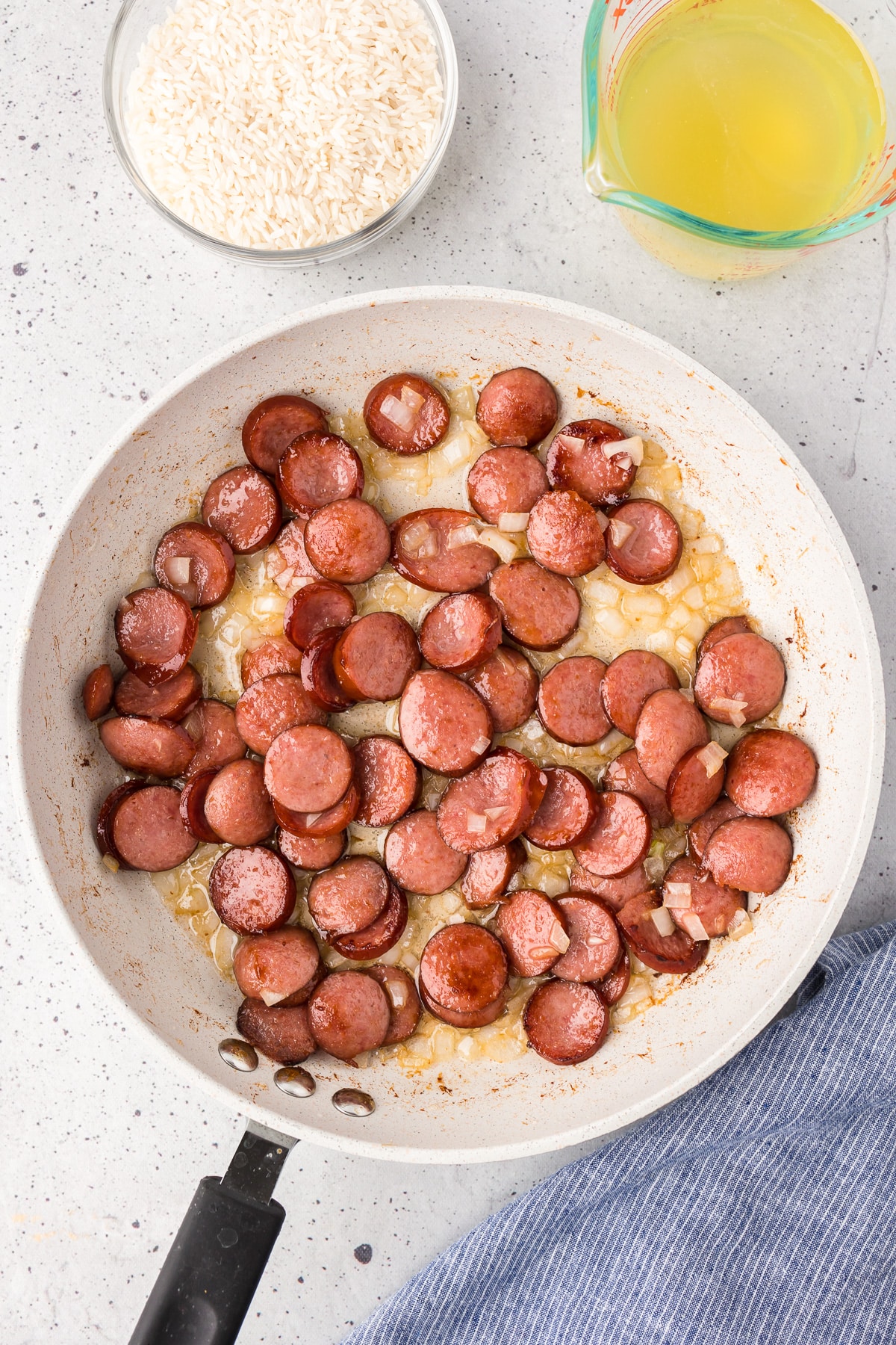 A skillet filled with cooked sausage pieces and onions, with a bowl of rice and a measuring cup of broth on the table nearby.