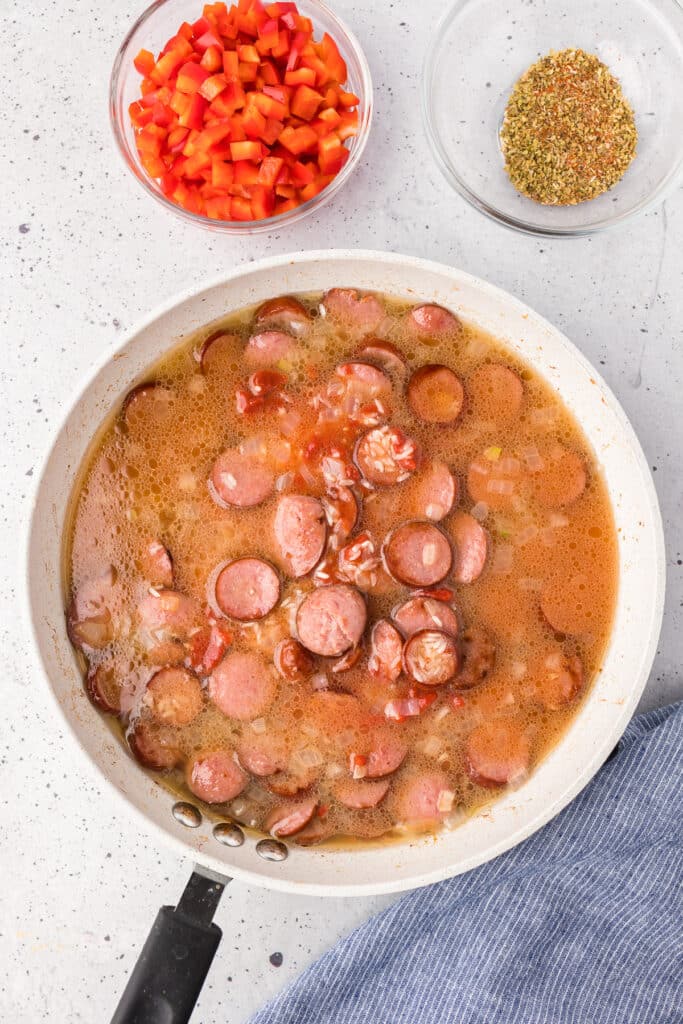 Sausage, onions and chicken broth in a skillet with peppers and spices in bowls nearby.