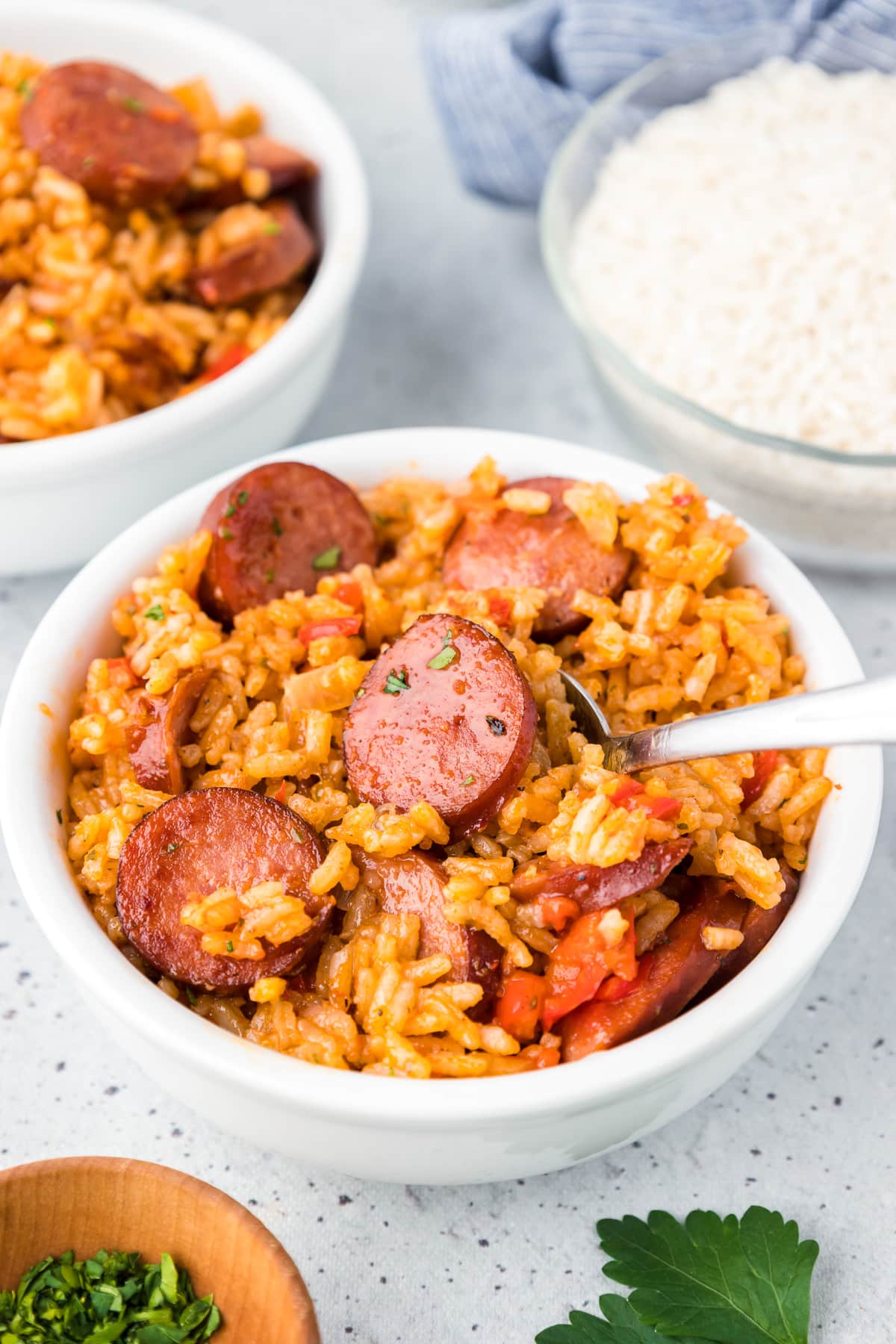 A bowl of rice and sausage with a spoon scooping with a family sized bowl full of more of the dish in the background.