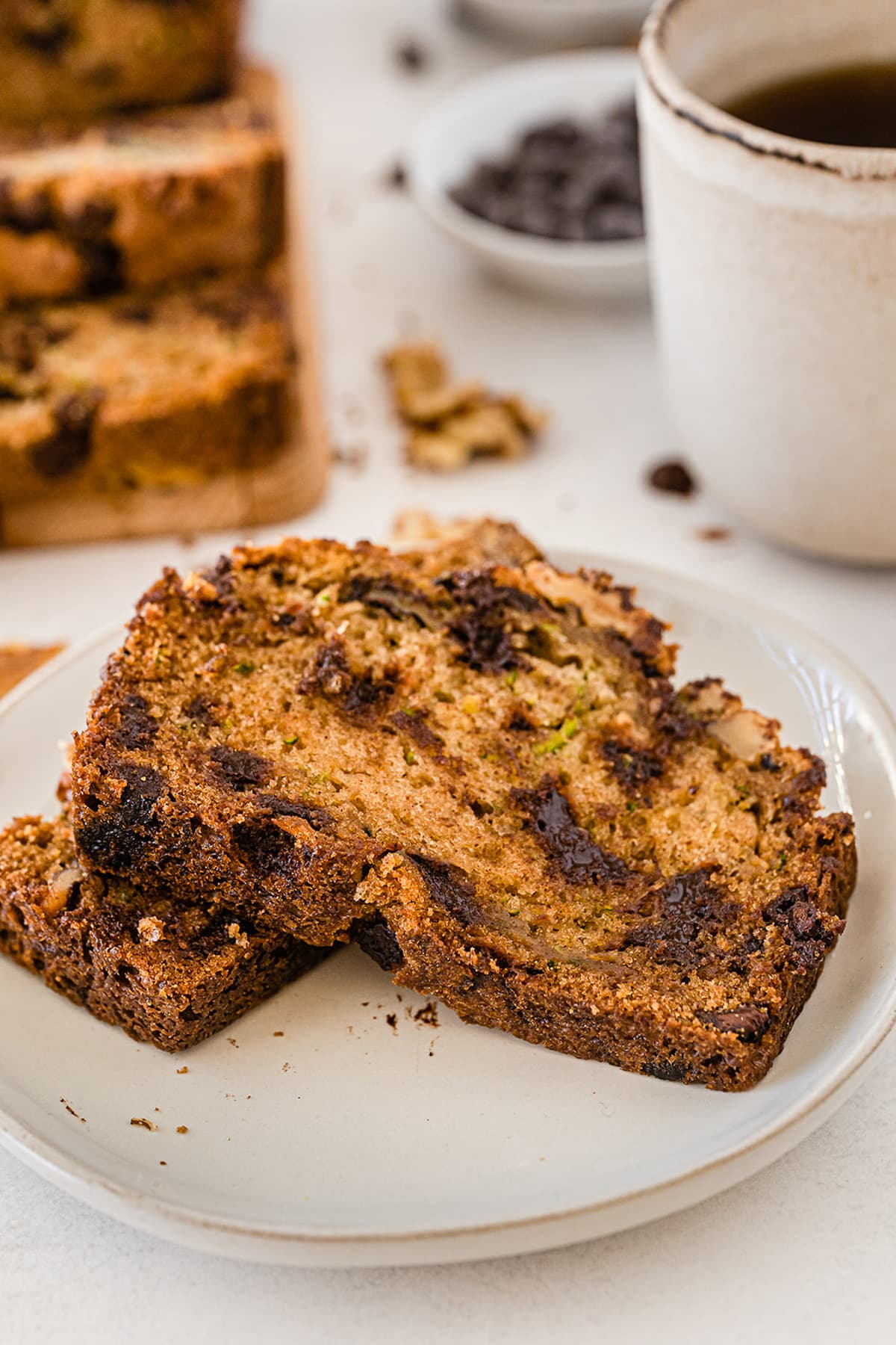 Two slices of chocolate chip zucchini bread with walnuts on a plate with the rest of the slices from the loaf and a cup of coffee in the background on the table.