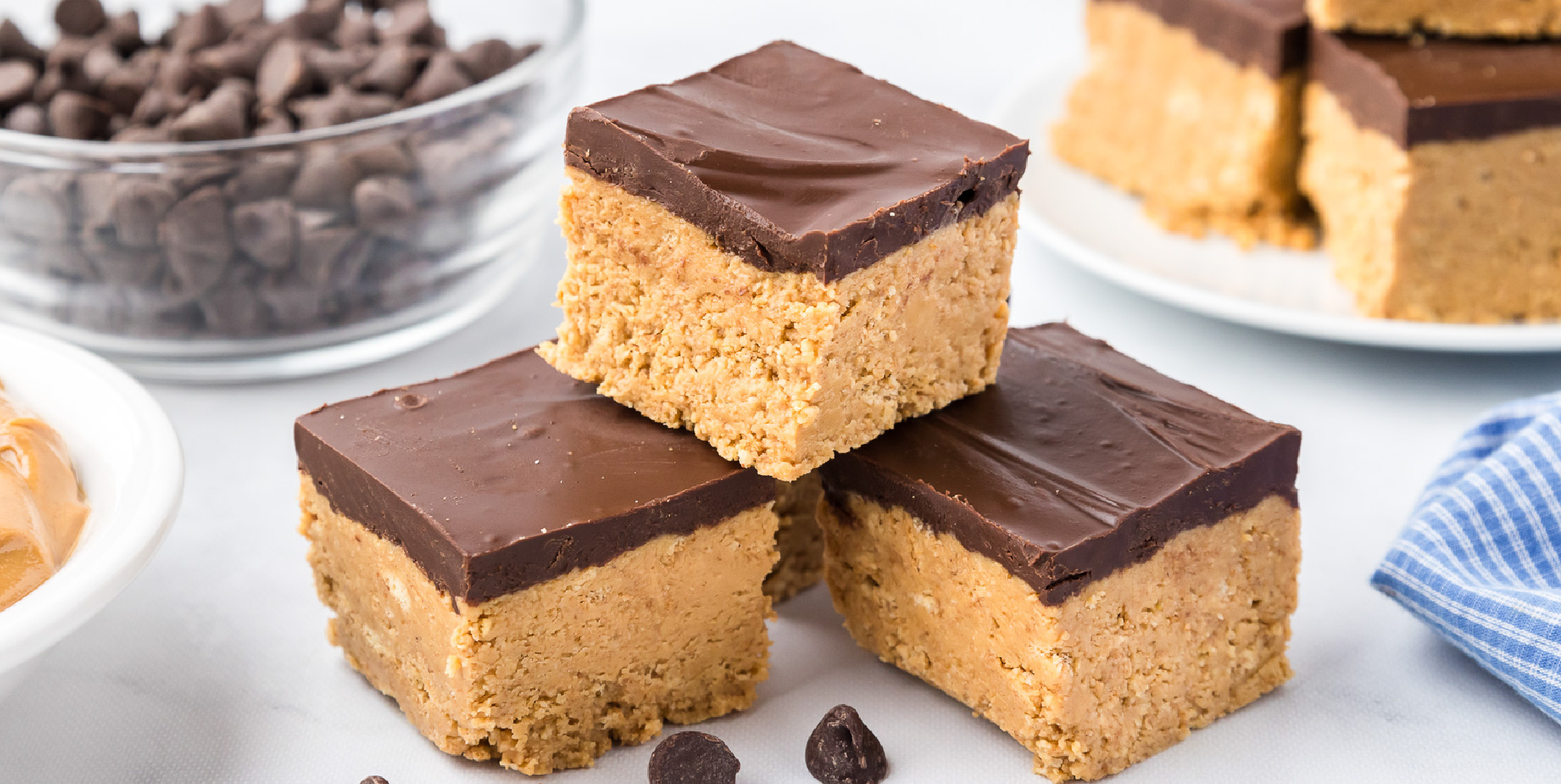 Wide view of stacked peanut butter squares on a counter with chocolate chips and more peanut butter squares in the background on the counter.