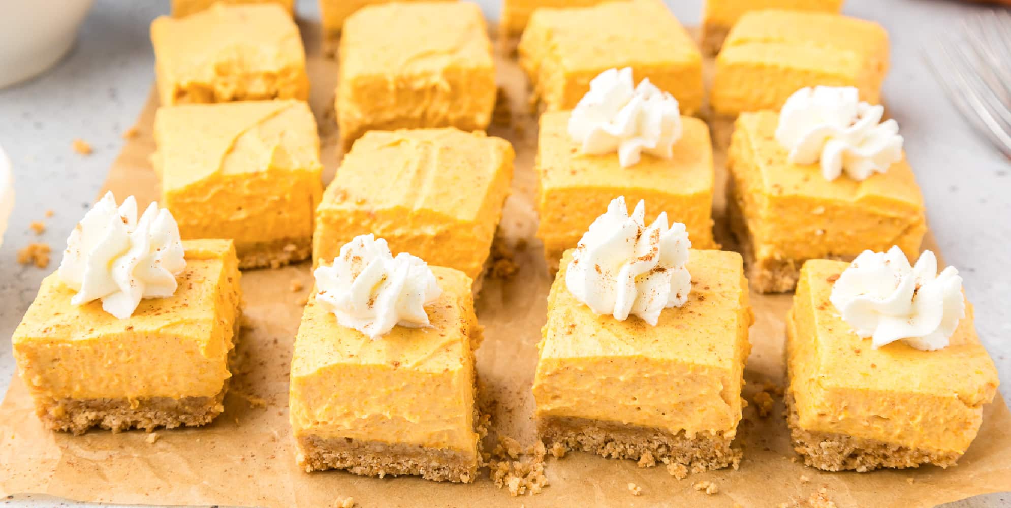 Wide view of a batch of pumpkin no bake cheesecake bars sliced on parchment paper from the side with some with swirls on whipped cream on top.