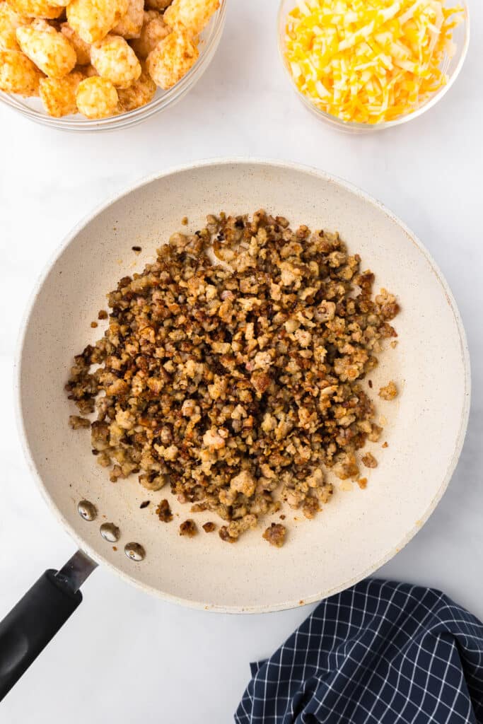 A frying pan filled with browned sausage and onions, with a bowl of tatter tots and cheese next to it on the pan.