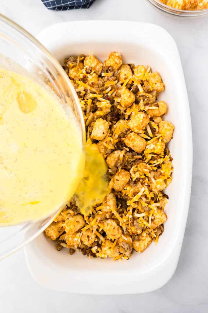 Egg mixture being poured from a bowl over tater tots, sausage and cheese mixed in a casserole dish.