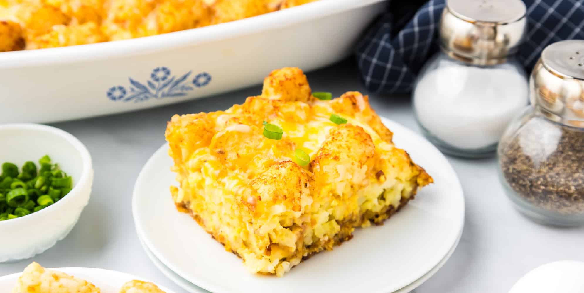 Cheesy tater tot casserole piece on a plate next to the casserole pan, and salt and pepper shakers.