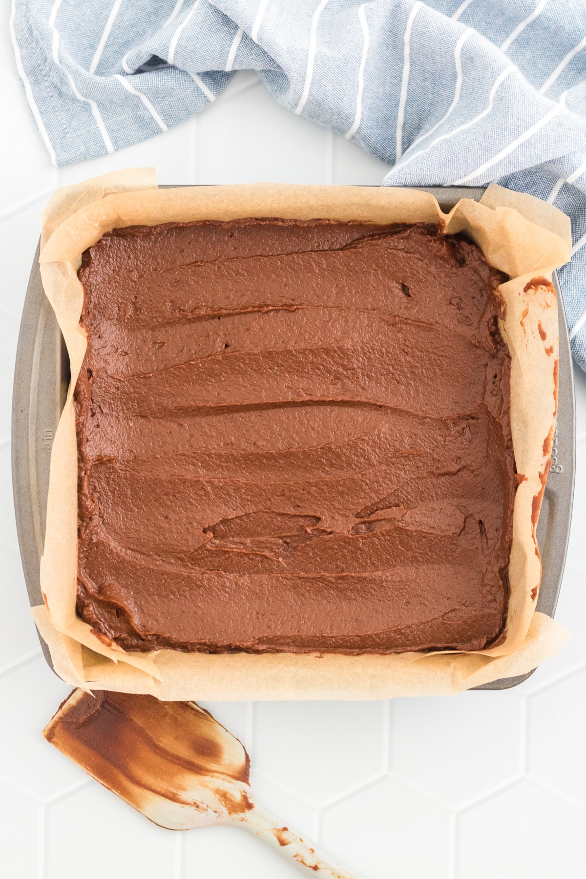 A chocolate pumpkin brownie dough smoothed in a parchment paper lined baking pan with a spatula on the counter nearby.