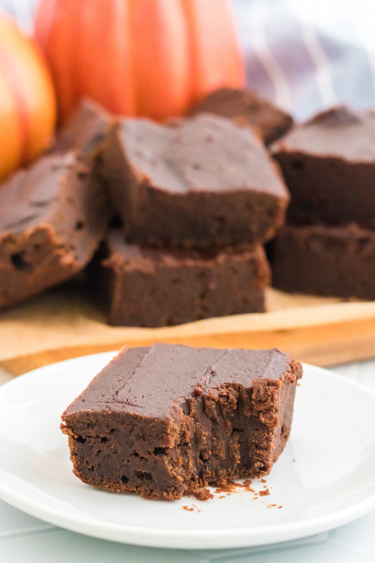 Chocolate pumpkin brownie on a white plate missing a bite with pumpkins and more stacked brownies in the background.