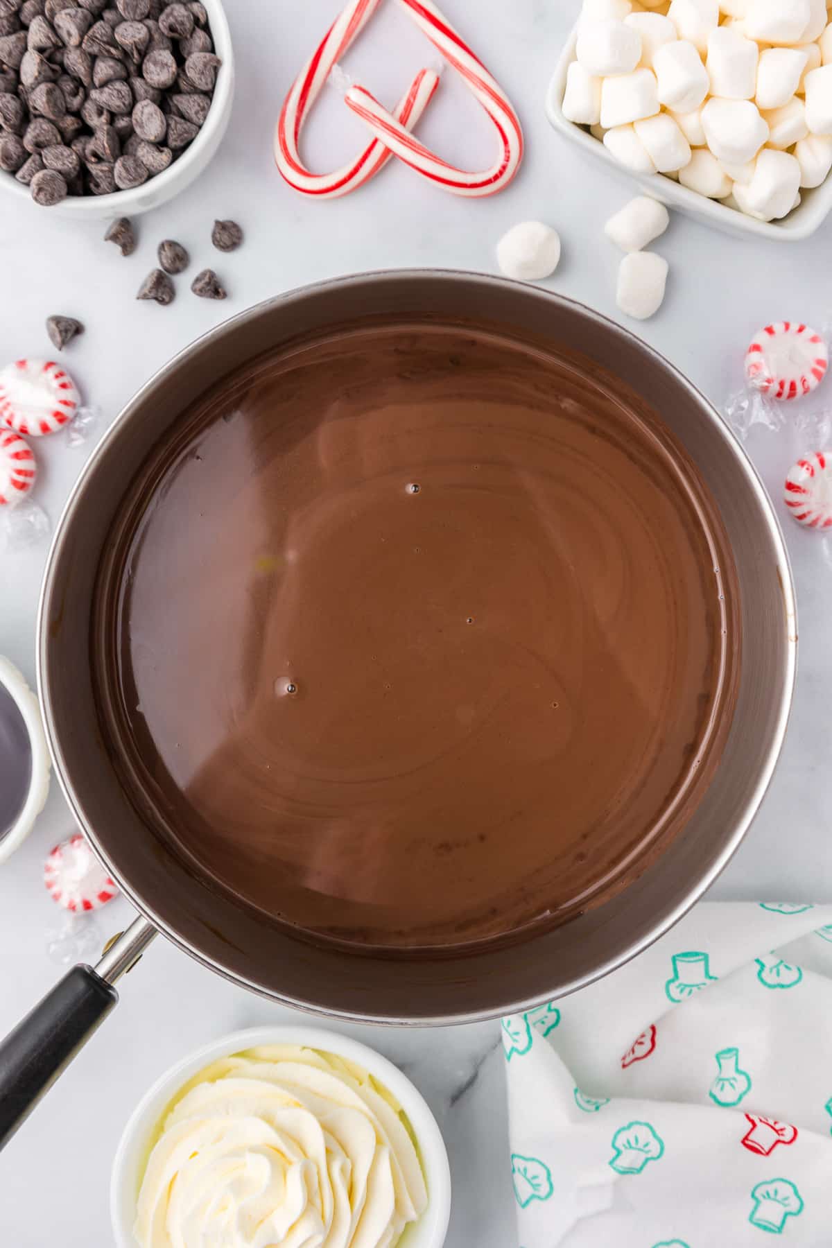 Hot chocolate in a pan with candy canes, whipped cream and marshmallows on the counter nearby.
