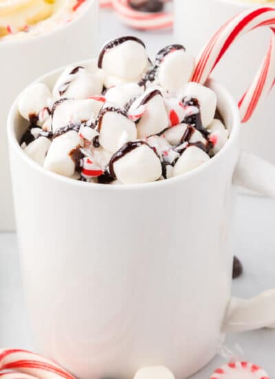 Hot chocolate with marshmallows and candy canes in a mug with more in the background.