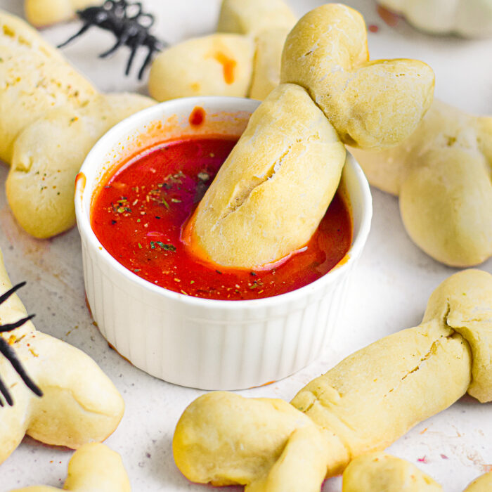 A bone shaped breadstick being dipped in a bowl with a red marinara sauce with more breadsticks nearby on the platter.