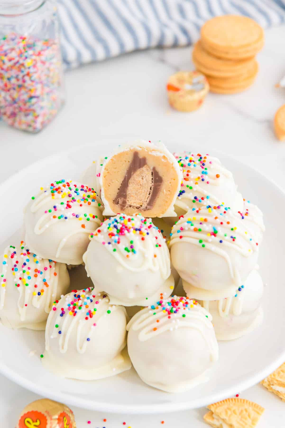 A plate of white chocolate truffles topped with sprinkles with a bite taken out of the top one showing a peanut butter cup inside.