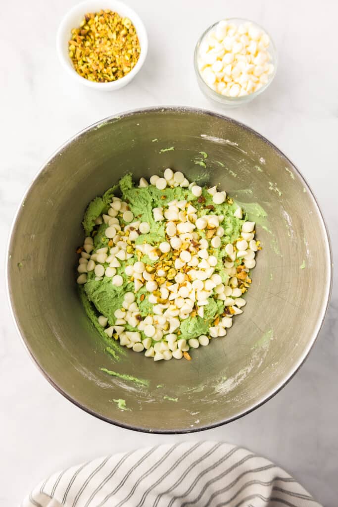 Green pistachio cookie dough in a mixing bowl from overhead with pistachio pieces and white chocolate chips on top of the dough to be mixed in.