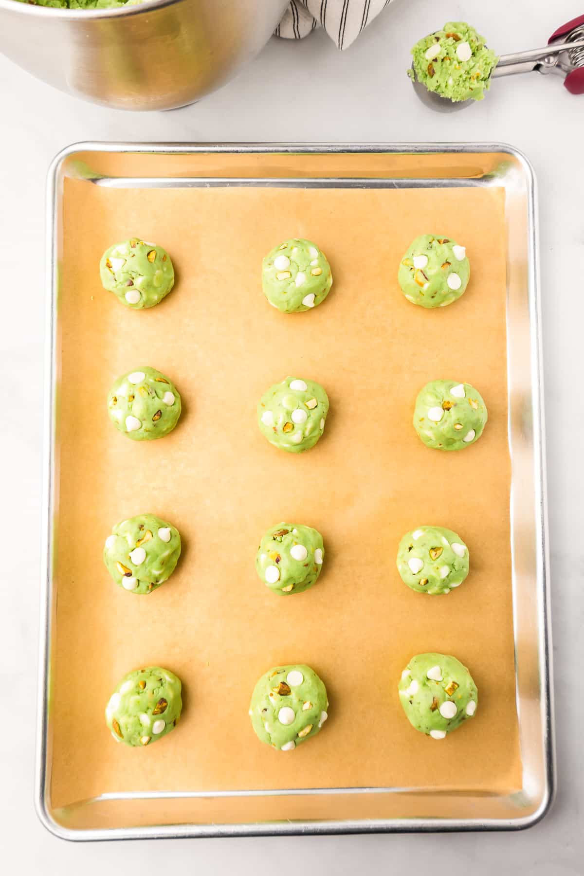 A baking sheet with green pistachio cookie dough topped with white chocolate chips on a baking sheet from above.
