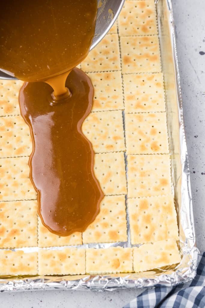 Liquid toffee being poured over saltine crackers on a baking sheet.