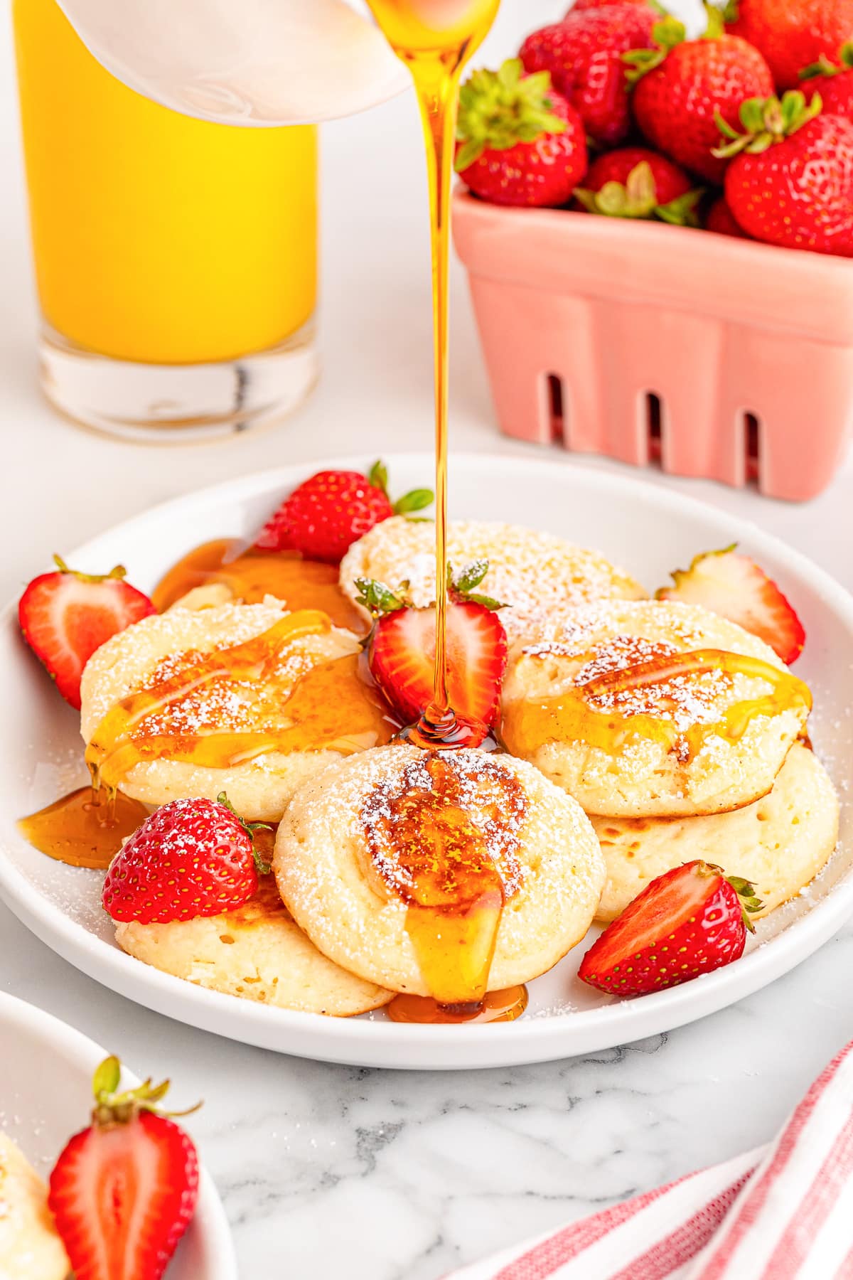 A plate of silver dollar pancakes topped with fresh strawberries with maple syrup being poured over them.