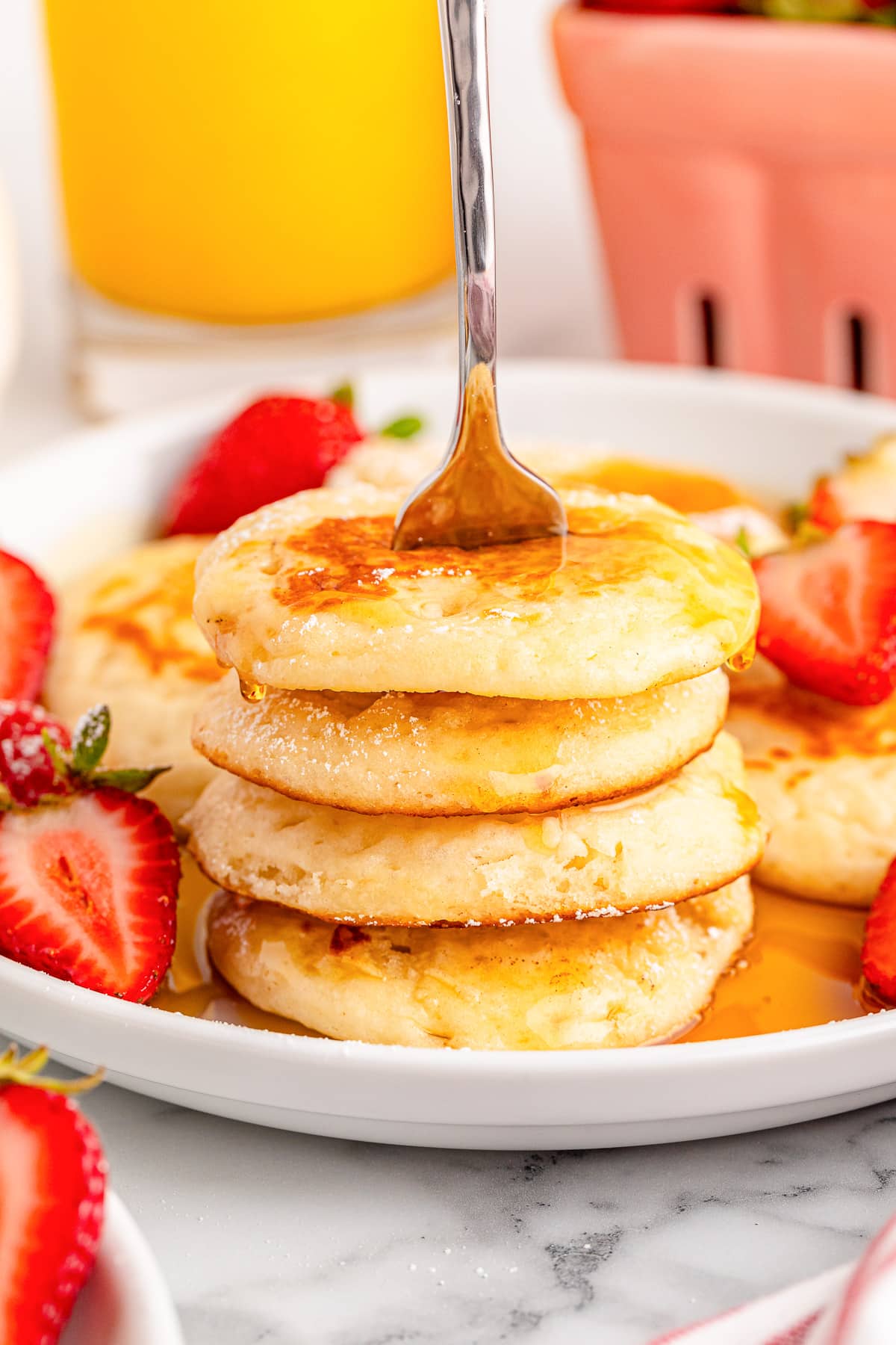 A stack of silver dollar pancakes being pokes with a fork with maple syrup and fresh strawberries on a plate from the side.