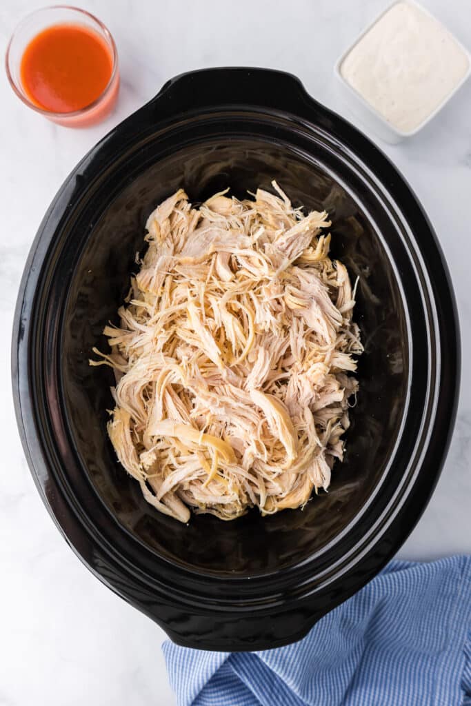 Shredded chicken in a crock pot with ranch dressing and hot sauce in small bowls nearby on the counter.