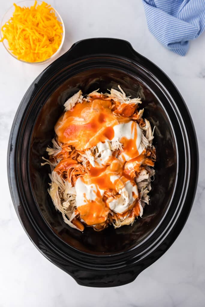 A crock pot filled with shredded chicken, ranch dressing and hot sauce with shredded cheddar cheese in a bowl nearby on the counter.