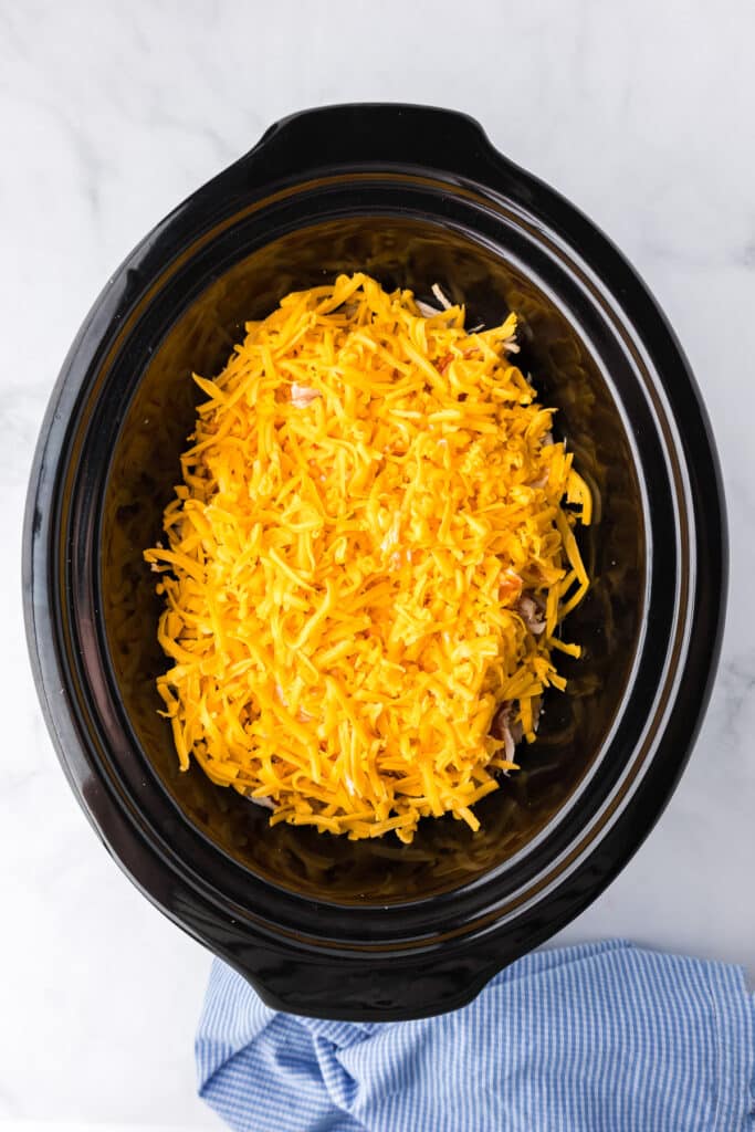 A crock pot filled with shredded cheese over chicken on a counter.