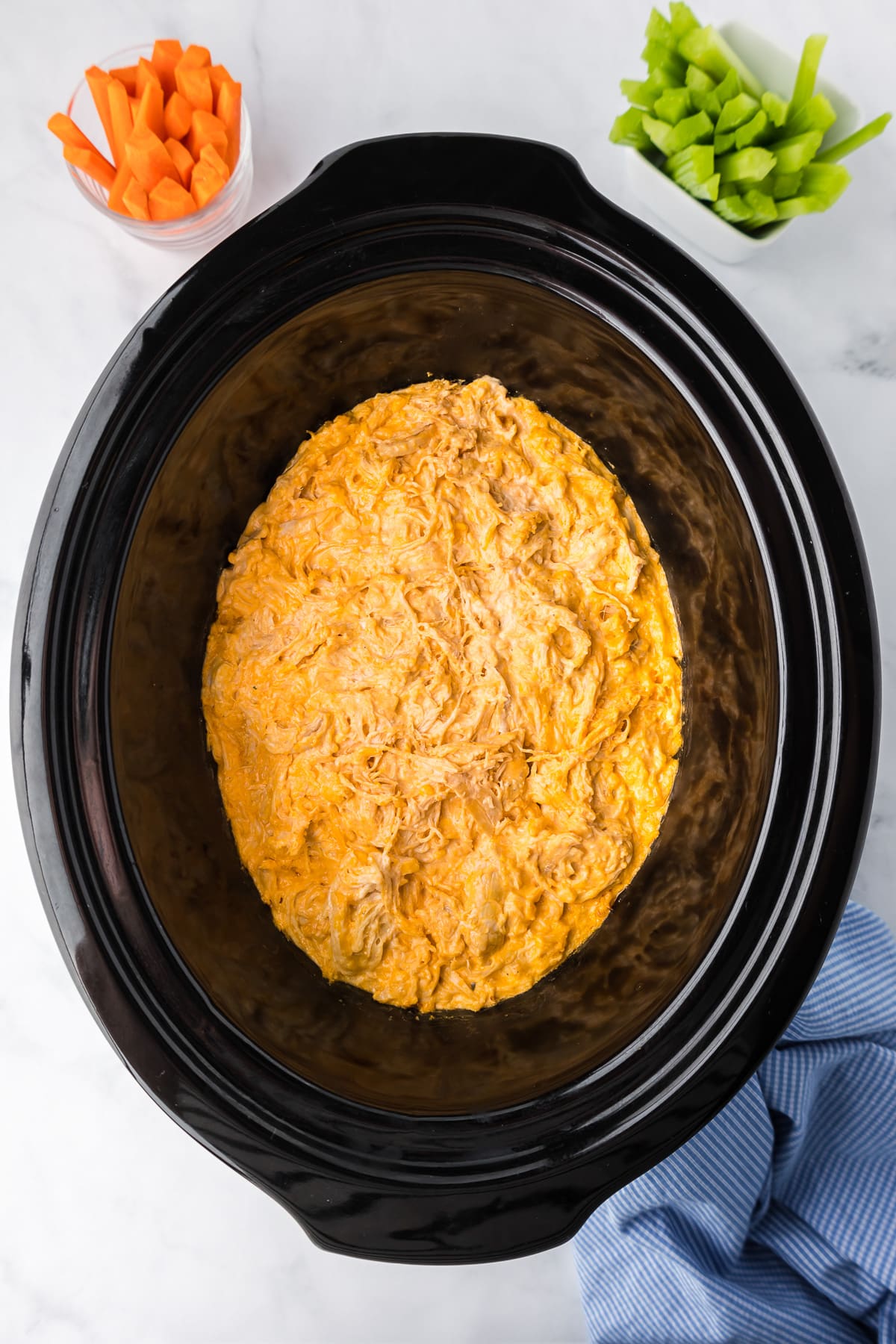 A crock pot filled with buffalo chicken dip with small cups of carrot and celery sticks nearby.