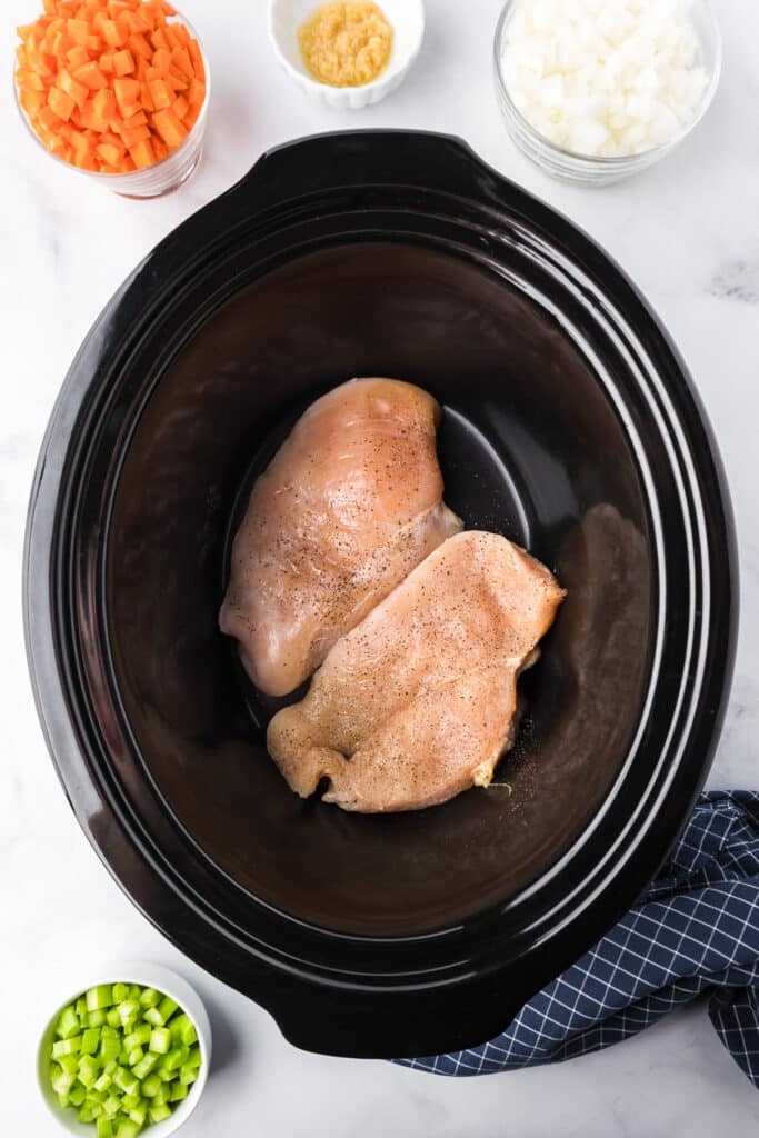 Chicken breast in the base of a slow cooker with bowls of diced carrots, celery and onions nearby.