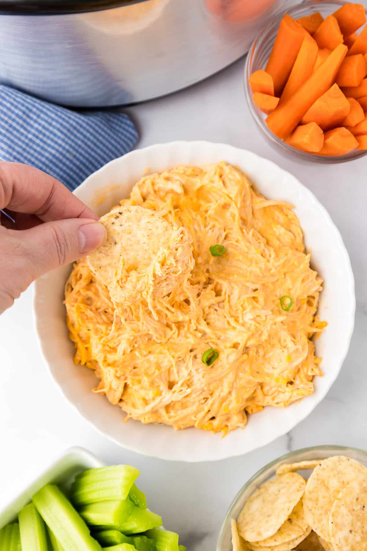 A person's hand dipping a chip into a bowl of buffalo chicken dip with more chips, carrots and celery in bowls nearby.