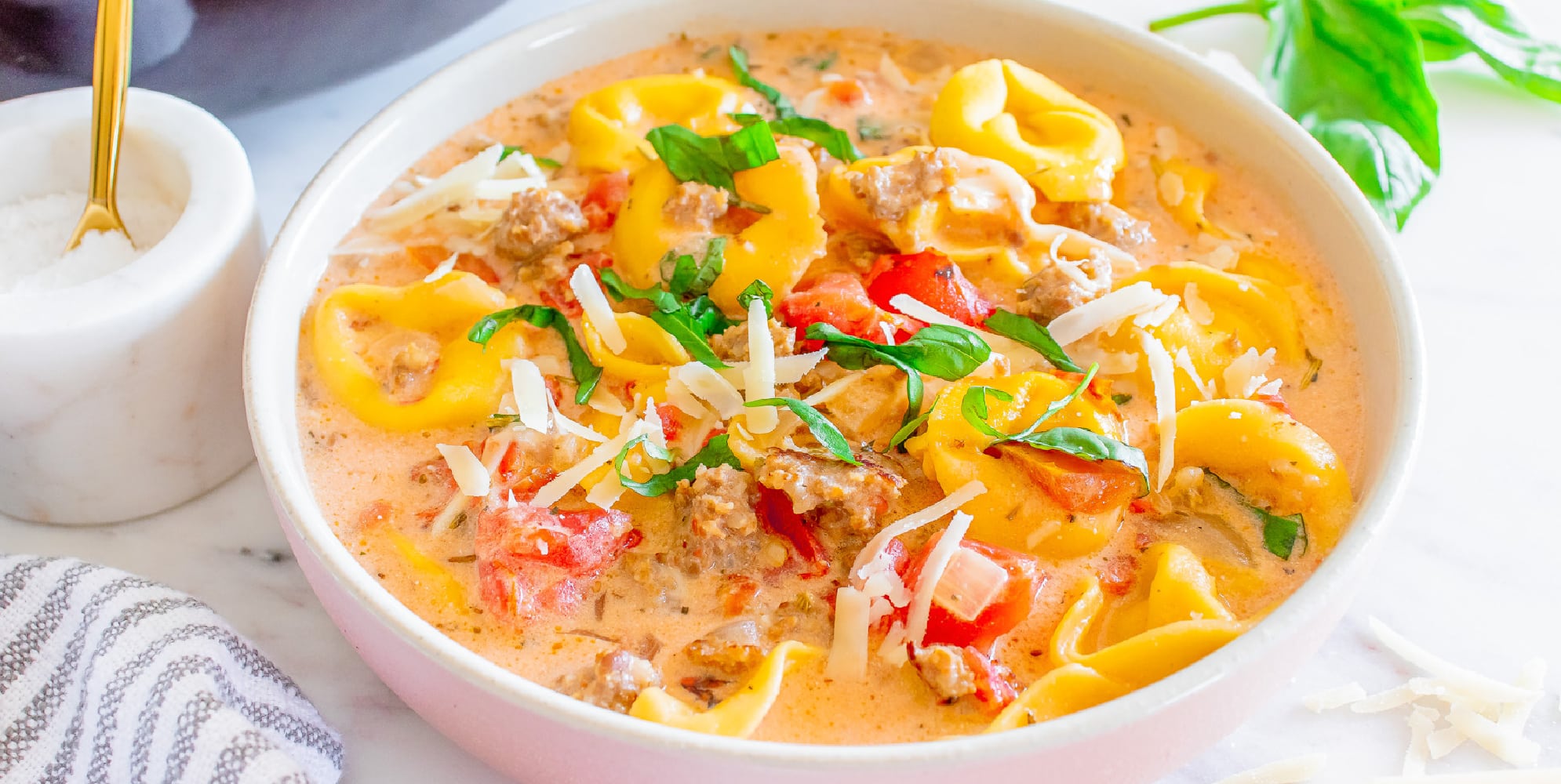 Wide view of creamy sausage tortellini soup in a white bowl from the side.