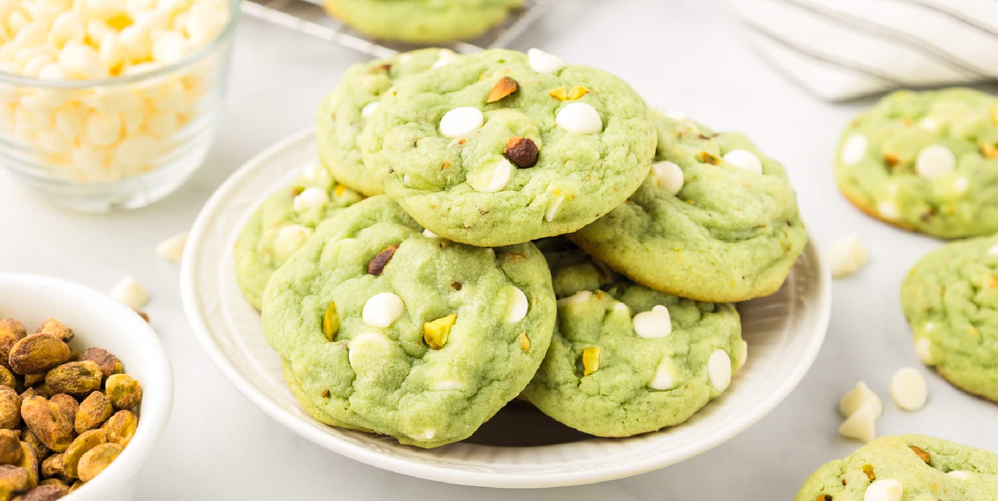 A wide view of a plate of green pistachio pudding cookies with white chocolate chips and nuts on top.