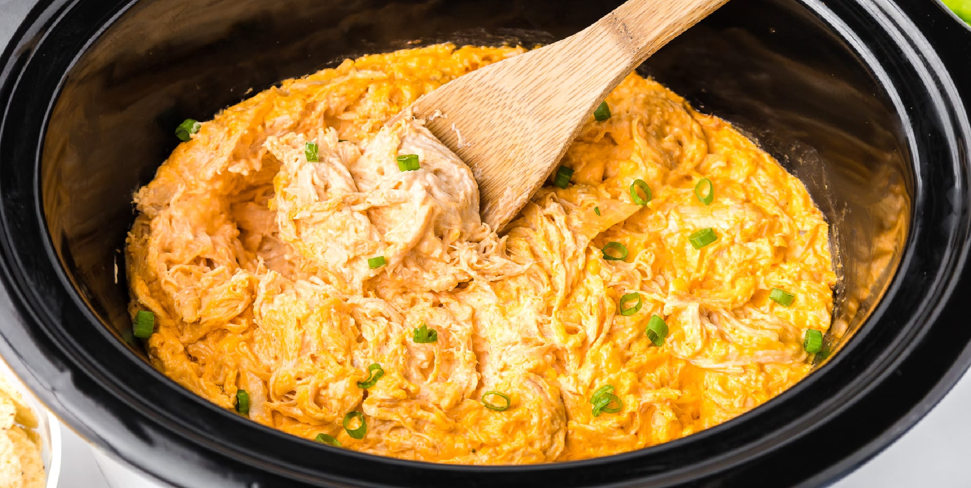 A crock pot full of chicken dip with a wooden spoon stirring it.