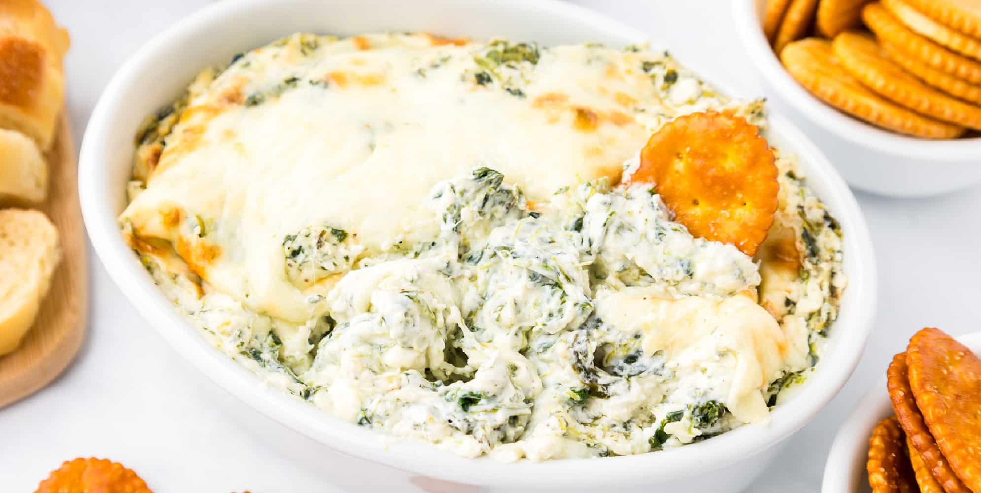 A bowl of spinach dip topped with melted cheese and with crackers being dipped in the dip.