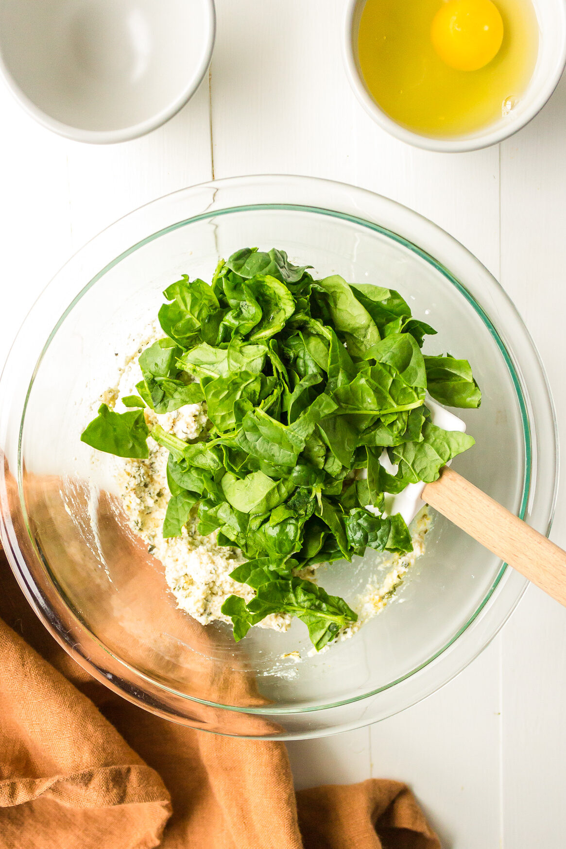 A large glass mixing bowl filled with spinach being mixed into a ricotta mixture with an egg in a bowl nearby.