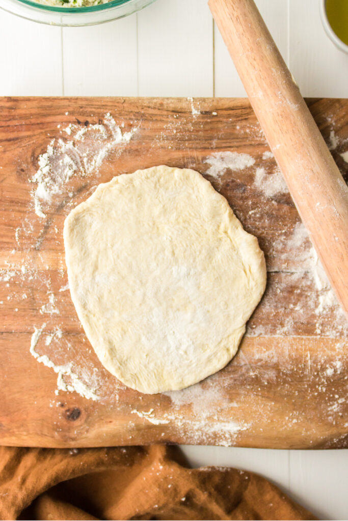 Pizza dough rolled flat on a wooden cutting board with a rolling pin nearby.