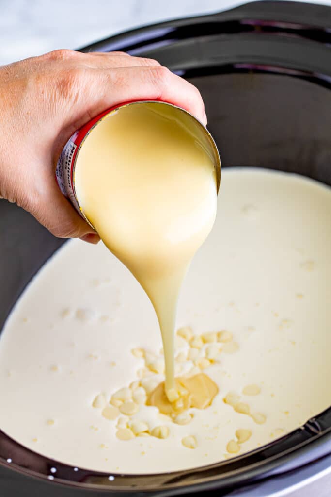 A person pouring sweetened condensed milk from a can into a slow cooker full of milk and cream.