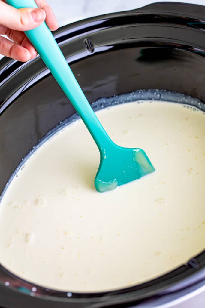 A person's hand stirring white hot chocolate in a slow cooker with a silicone spatula.
