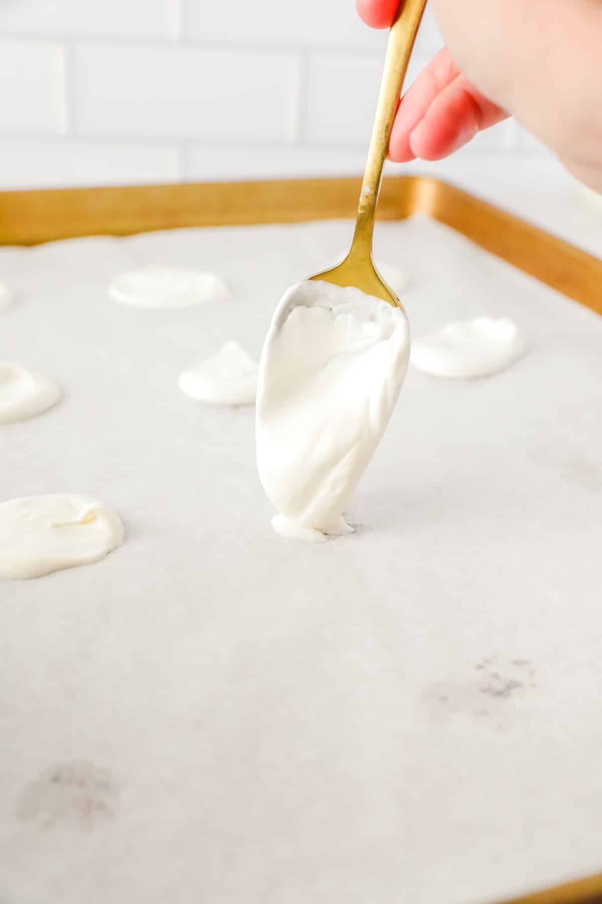 A person's hand using a spoon to spread dollops of white candy melts on a baking sheet.