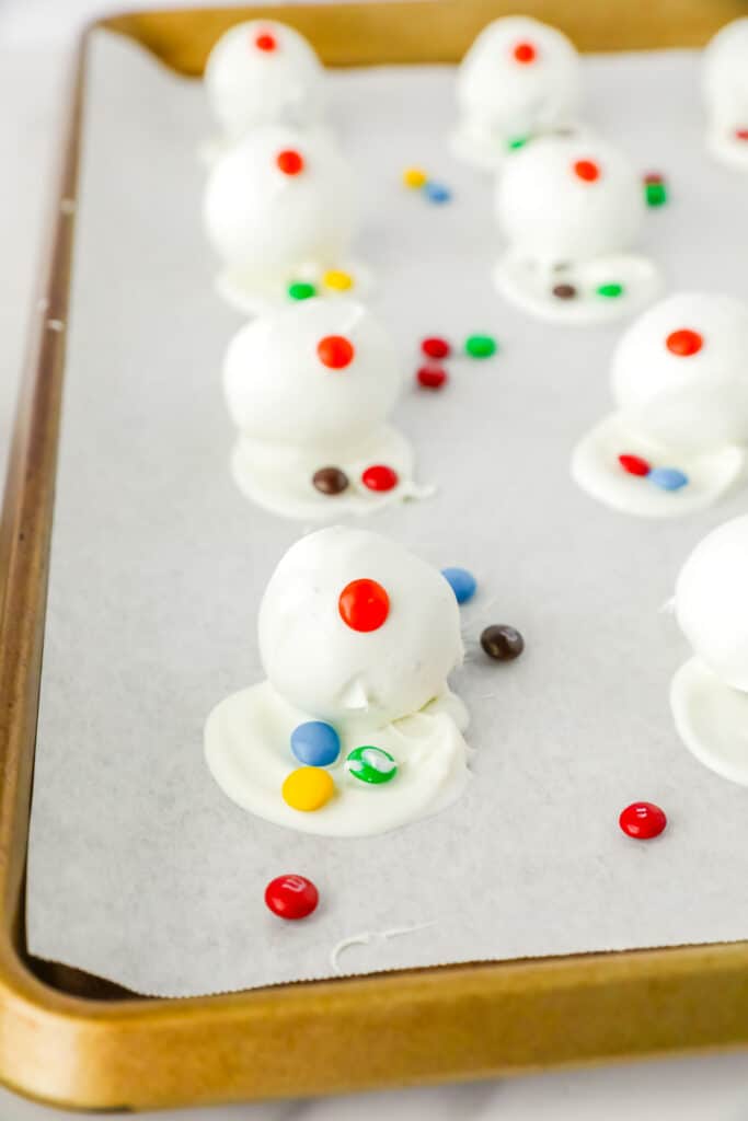 Snowman oreo balls on a baking sheet being decorated with mini M&Ms.