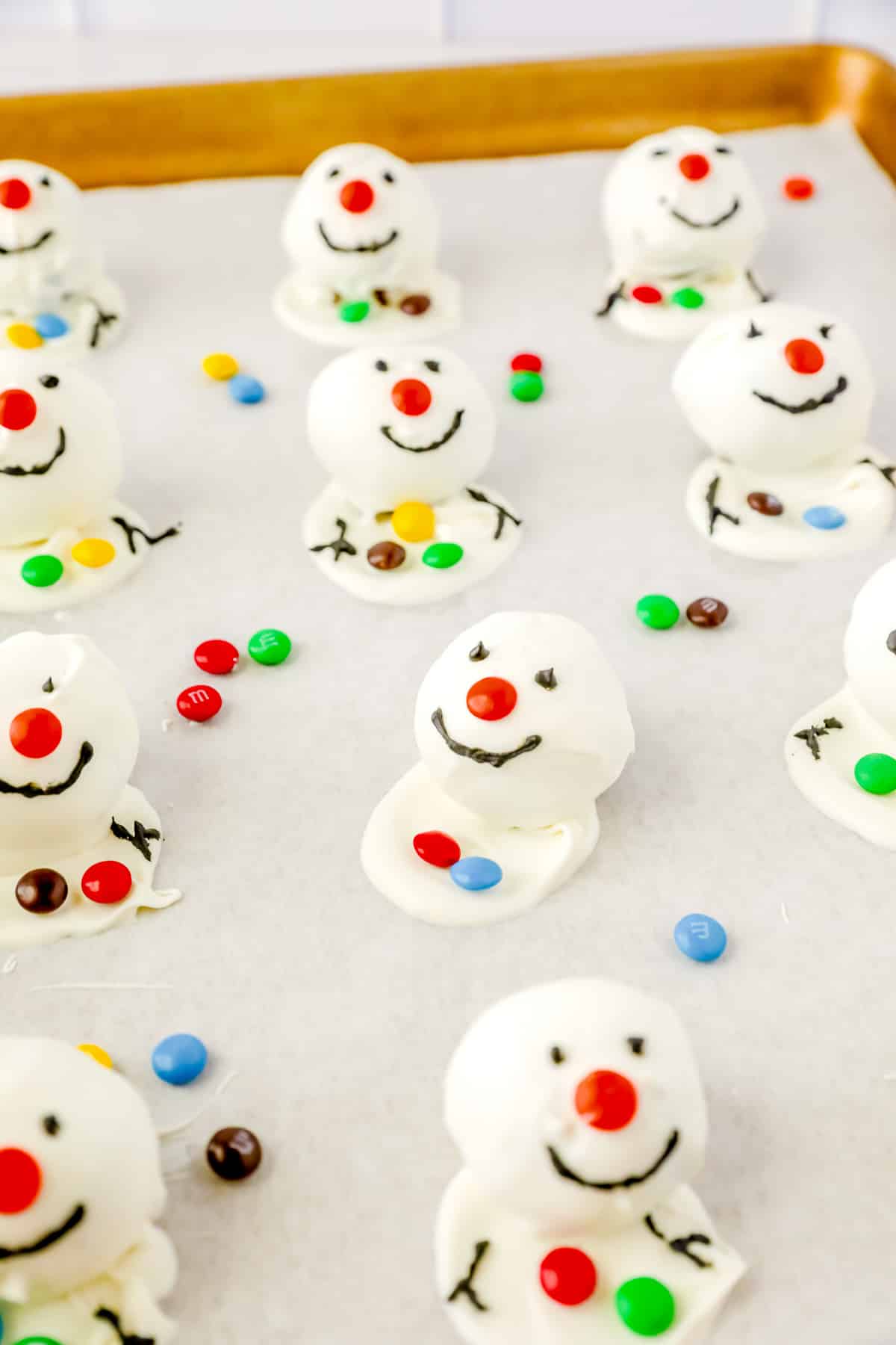 Snowman oreo balls s on a baking sheet decorated with eyes, smiles and stick arms with black decorating gel.