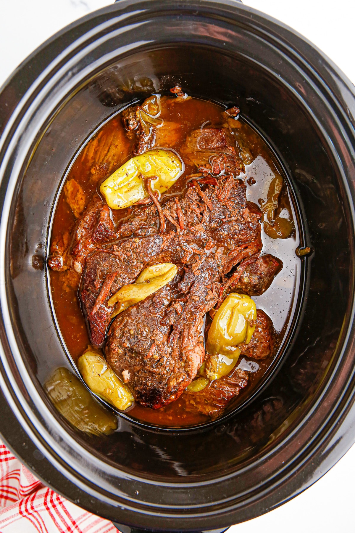 A crock pot full of a large piece of mississippi pot roast chuck roast after cooking sitting in juice topped with pepperoncini peppers.