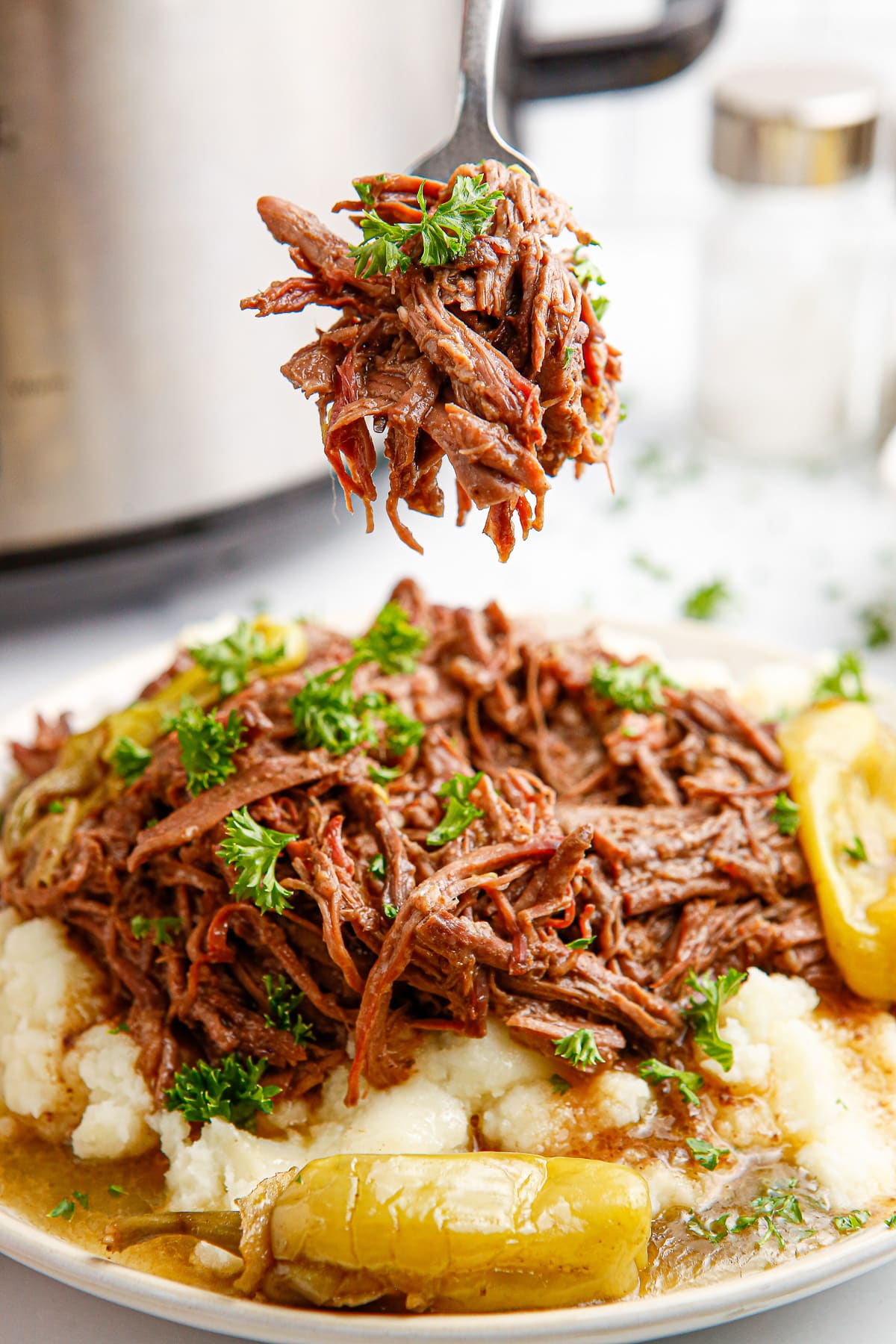 A bite of slow cooker Mississippi pot roast being lifted from a plate of shredded beef on top of mashed potatoes by a fork.