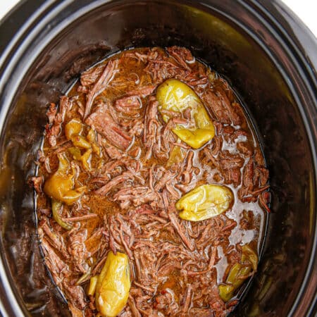 A slow cooker filled with shredded Mississippi pot roast beef, cooking liquid and peppers from above.