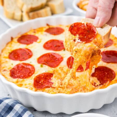 A person dipping into a round pie pan of pepperoni dip with a piece of bread stretching gooey cheese.